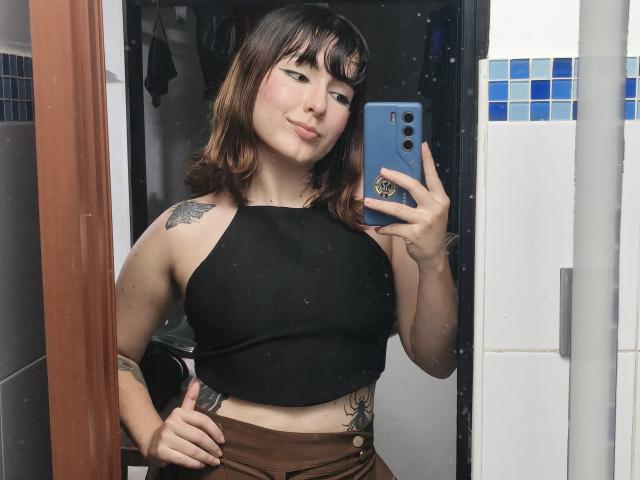 ValeriHairy69 with brown hair, black crop top, brown skirt, tattoos on shoulder and side, holding blue phone, mirror selfie, slight smirk, white and blue tiled bathroom.