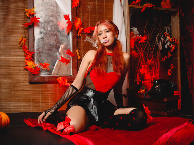 SuriEverly with red hair, wearing a red top, black fishnet arm sleeves, black leather skirt, and thigh-high socks, sitting on a red cloth, surrounded by autumn leaves.