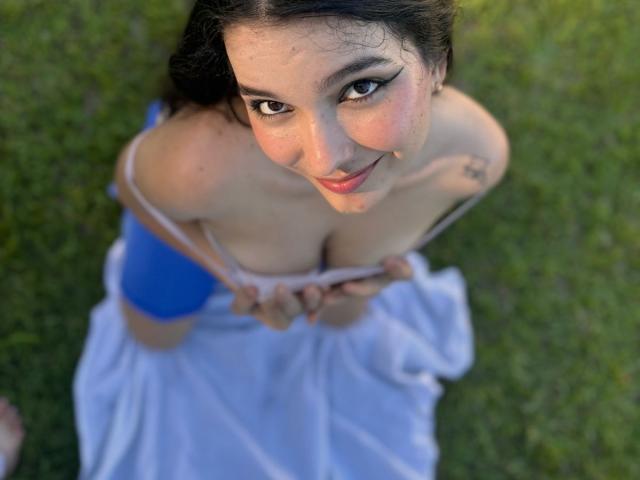 ValeriHairy69 with large tits, pulling down a pink top, wearing blue shorts, looking up with a smile, standing on grass, hands on her chest.