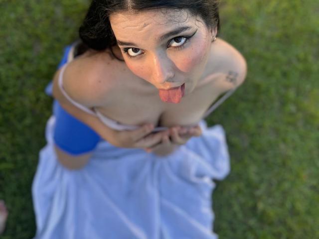 ValeriHairy69 with dark hair, pink eyeliner, and tongue out, kneeling on grass, holding her tits, wearing a blue top and white skirt, looking up.