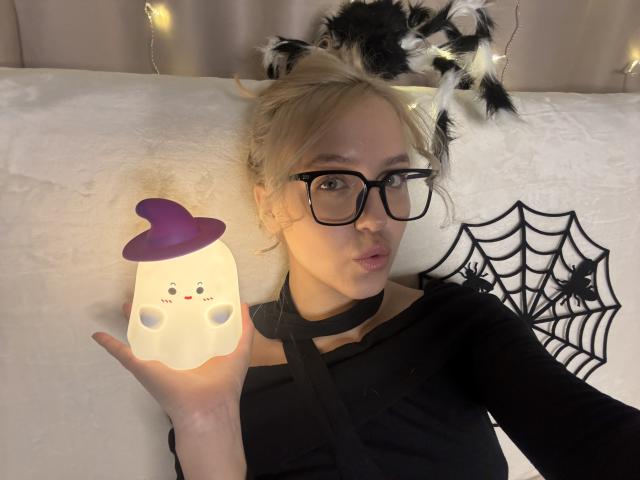 CindyLu met blond haar, zwarte bril en een zwarte choker, die een lantaarn in de vorm van een spook vasthoudt met een paarse hoed, draagt een zwarte top en ligt op een wit bed met spinnen decoratie..