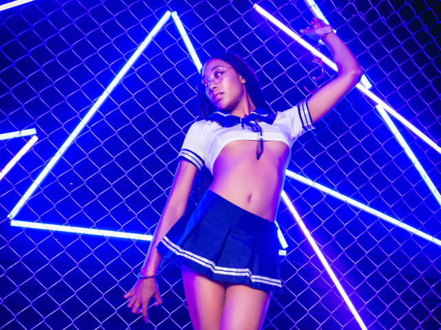 EsmeraldaRoe in a schoolgirl outfit, white crop top, navy mini-skirt, exposing her tits, standing against neon triangles, arms raised, looking up, blue-purple neon lighting.