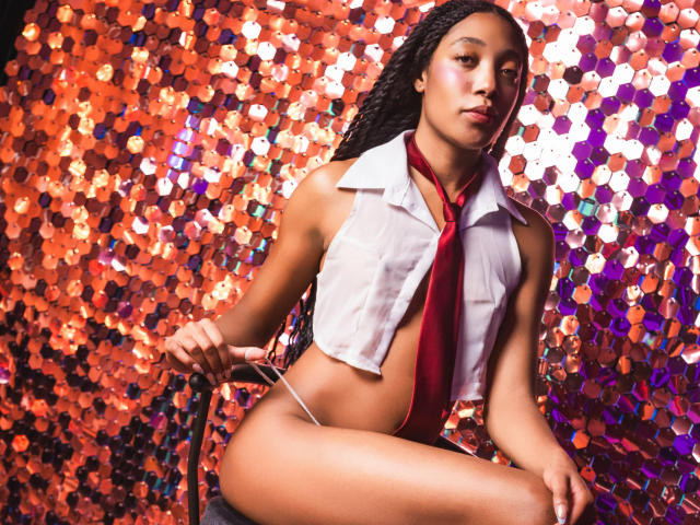 EsmeraldaRoe with long braids, wearing a white sleeveless shirt, red tie, and thong, sitting against a shiny orange and purple sequined background, looking at the camera.