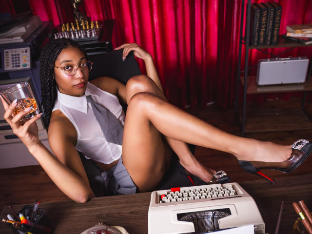 EsmeraldaRoe with dark skin, braided hair, glasses, white sleeveless shirt, grey tie, grey shorts, holding glass of whiskey, legs up, black heels, typewriter, and ammo in front.