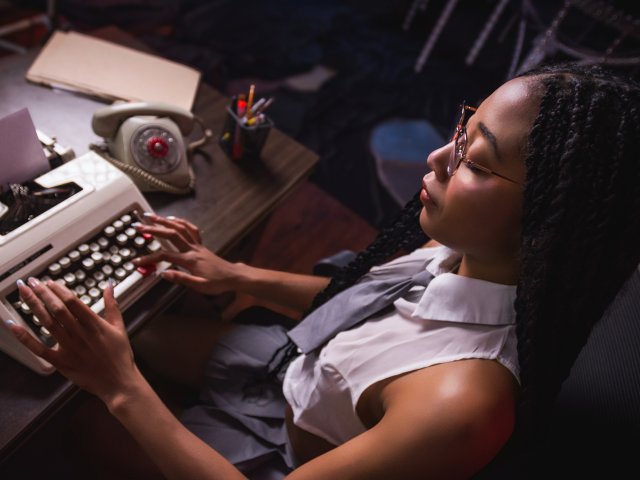 EsmeraldaRoe with braided hair, wearing glasses and a sleeveless white shirt, typing on an old-school typewriter, her hands on the keys, in a dimly lit room.