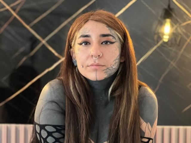DacotaSexy, con lunghi capelli castani, piercing al naso, tatuaggi sul viso e una maglia a rete nera, seduta di fronte a un muro con motivo a rombi, con una lampadina sullo sfondo..