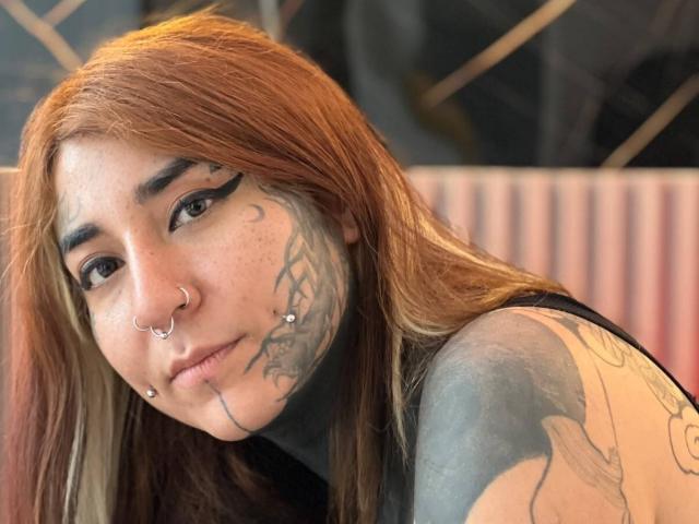 DacotaSexy, com cabelo castanho-avermelhado, piercing no nariz, piercing no lábio, tatuagens no rosto e no braço, blusa de gola alta preta, olhando para a câmera, expressão suave, sardas claras, maquiagem discreta, ambiente interno..