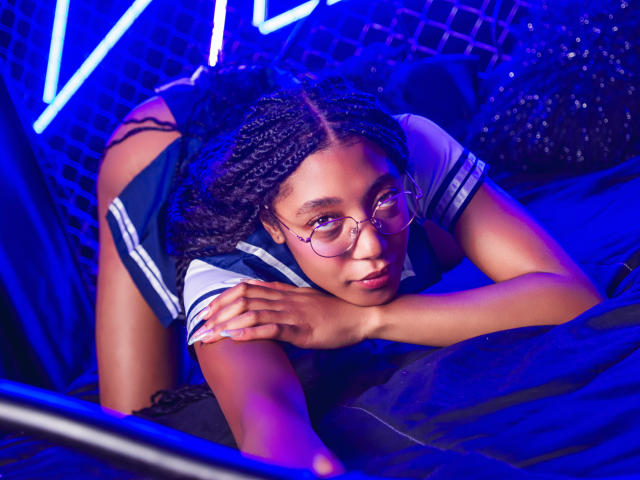 EsmeraldaRoe on all fours, wearing a navy and white schoolgirl outfit, with glasses, and her braided hair, looking at the camera, with blue neon lights in the background.