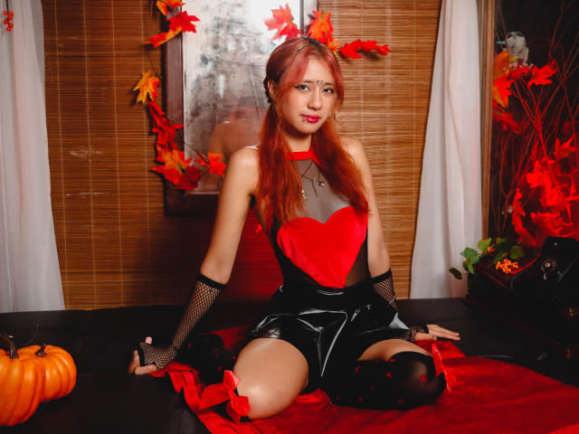 SuriEverly with red hair, wearing black fishnet gloves, red heart top, black shiny shorts, and red thigh-highs, kneeling on red fabric, surrounded by pumpkins and autumn leaves.