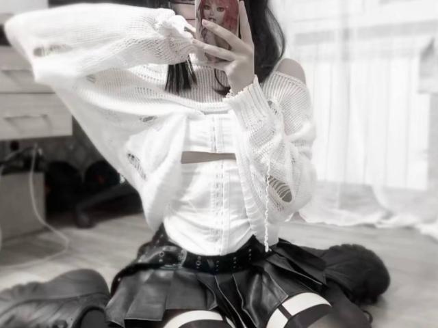 JaclynJafek with white knit sweater, black pleated skirt, white crop top, black belt, and garter straps, taking a selfie with phone, showing her tits and ass.