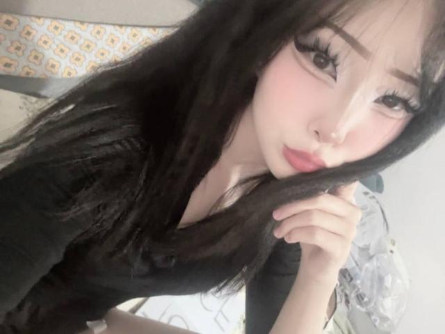 JaclynJafek with long black hair, pink makeup, and black top, touching her chin, looking at the camera with half-closed eyes, showing her pink lips and big eyelashes.