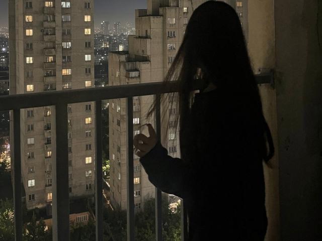 Silhouetted JaclynJafek with long dark hair stands on a balcony, holding a phone, looking at tall buildings with lit windows at night. She's in a dark, partially visible outfit.