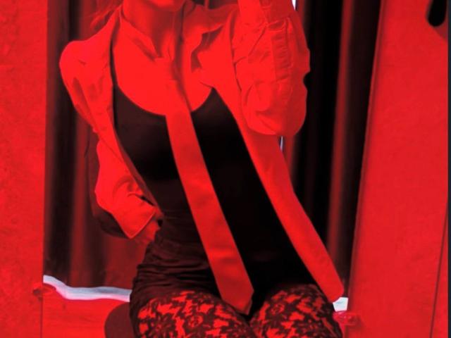 JaclynJafek in a red shirt with a black tank top, red tie, and lace leggings, sitting with hands on her hips, red lighting, showing her ass.