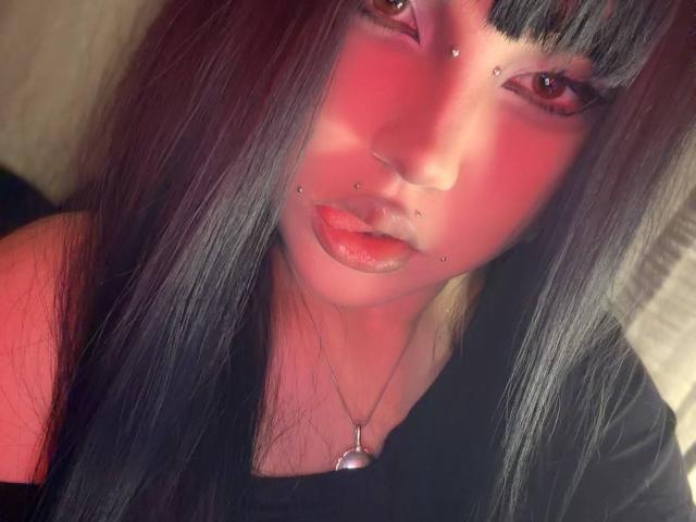 AlmaStampka has black straight hair, pinkish-red makeup, and multiple facial piercings including her nose and lower lip. She wears a black shirt and a silver necklace.