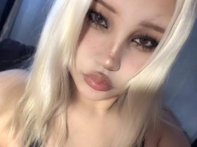 AlmaStampka, met platinablond haar, volle roze lippen en grote bruine ogen, kijkt in de camera met een lichtelijk gekanteld hoofd. Haar kleine borsten zijn nauwelijks zichtbaar..