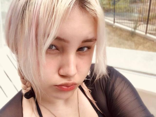 AlmaStampka, con i capelli rosa pallido e biondo platino, le labbra rosa carnose e una maglietta nera trasparente, guarda in basso con un'espressione leggermente infastidita. I suoi occhi azzurri sono socchiusi..
