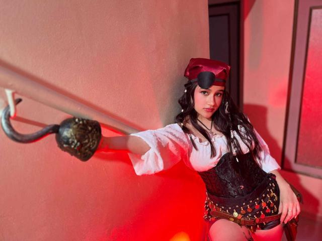 IrisBlair with long black hair, wearing a red beret, white puffed shirt, black corset, and short skirt with belt, leaning on a brass-handled pole, in red lighting.