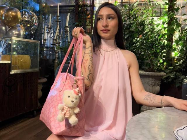 MaddyCarina with long dark hair, pink halter dress, holding pink bag with teddy bear, tattoos on arm, sitting at marble table, looking up with slight smile.