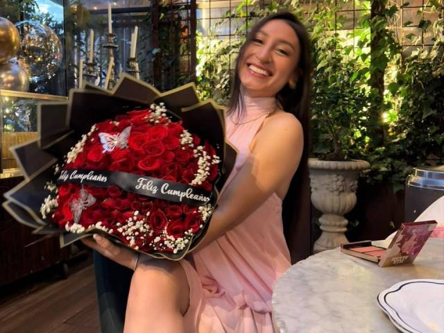 MaddyCarina with a big ass bouquet of red roses and white flowers, wearing a pink dress, smiling, holding the bouquet in a restaurant with green plants.