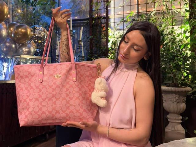 MaddyCarina with long black hair, wearing a pink dress, holding a large pink Chanel handbag with a fluffy keychain, her tits visible through the fabric.