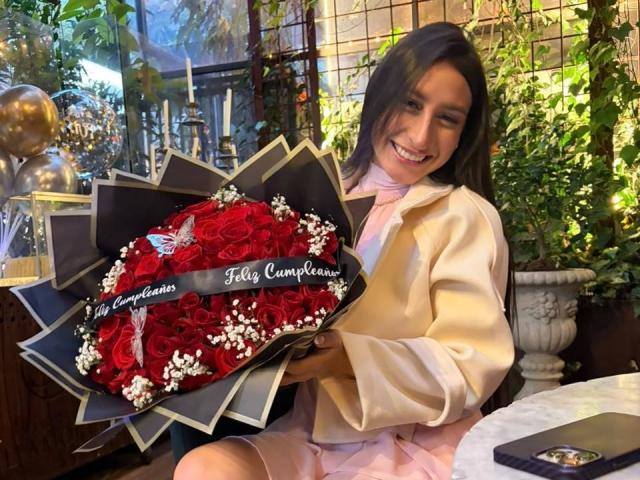 MaddyCarina with a smile, holding a heart-shaped bouquet of red roses and white baby's breath, wearing a pink dress and white jacket. Her ass is slightly visible.