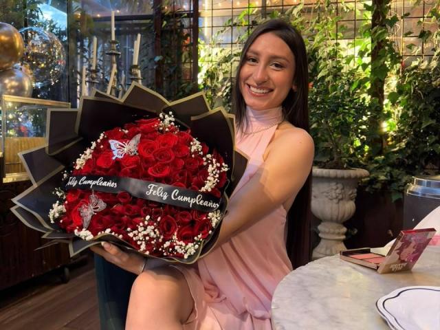 MaddyCarina with long brown hair, pink dress, holding red rose bouquet, white baby's breath, black and gold wrapping, smiling at camera, sitting at white marble table.