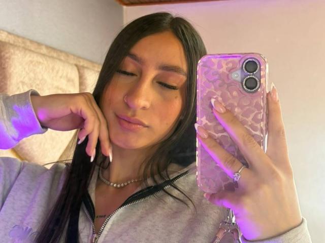 MaddyCarina with long brown hair, closed eyes, and a slight smile, holding a pink leopard-print phone, wearing a gray zip-up hoodie, and a silver necklace.
