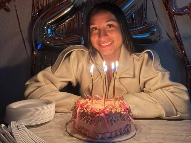MaddyCarina with straight brown hair, smiling, wearing a beige coat, sitting at a table with a pink frosted cake and seven lit candles, white plates in the foreground.
