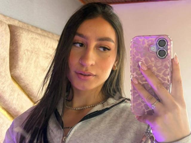 MaddyCarina with long brown hair, light skin, wearing a gray zip-up, holding a pink leopard phone case, pink-nosed, subtle makeup, necklace, and ring, taking a selfie.
