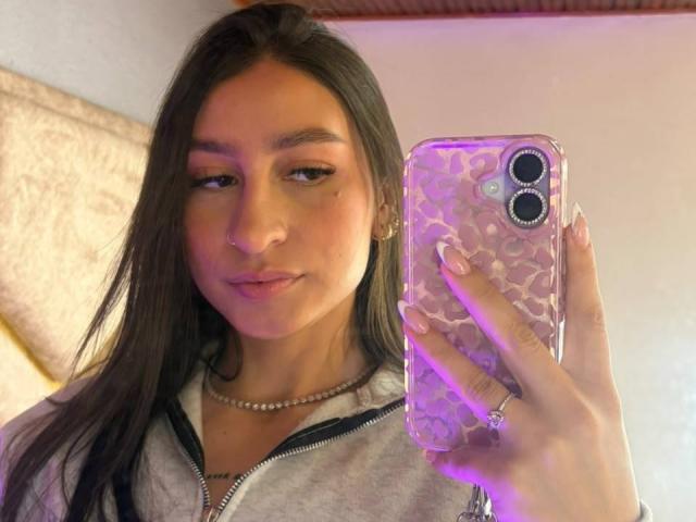 MaddyCarina with long brown hair, light brown skin, wearing a gray hoodie, holding a phone with a purple-patterned case, taking a selfie, showing a subtle smile, and a ring.
