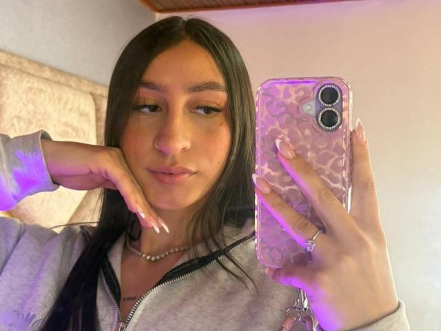 MaddyCarina with long black hair, light brown skin, and cute face, holding a phone with a pink patterned case, wearing a grey zip-up hoodie, and a ring on her finger.