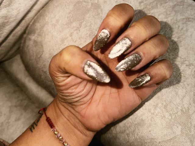 ThaliaAres' Hand mit glitzernden, silberfarben lackierten Nägeln, die funkelnde, metallische Spitzen an jedem Finger zeigen. Sie trägt ein rotes Perlenarmband an ihrem Handgelenk..