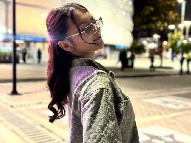 AlessiaFerry, con lunghi capelli scuri ondulati raccolti in una coda di cavallo, indossa occhiali e una giacca verde scintillante, e guarda indietro con un leggero sorriso. Strada cittadina di notte, sfondo sfocato..