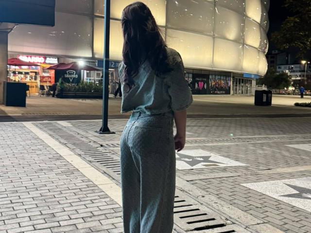 AlessiaFerry è di spalle rispetto alla fotocamera, indossa una maglietta grigia e pantaloni a vita alta, con i glutei leggermente in evidenza, in una scena notturna urbana poco illuminata..
