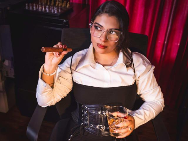 IsabellaFleur with glasses, holding a cigar in one hand and a whiskey glass in the other, wearing a white shirt, black corset, and red lipstick.
