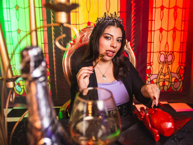 IsabellaFleur with a crown, sucking on a lollipop, holding a ring, wearing a black top, purple tank, and a necklace, with a bong, glass, and red crab in front.