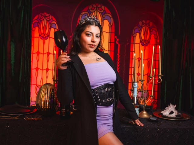 IsabellaFleur with dark hair, wearing a silver tiara, black coat, lavender dress, and belt, holding a black glass, standing in a dimly lit room with candles.