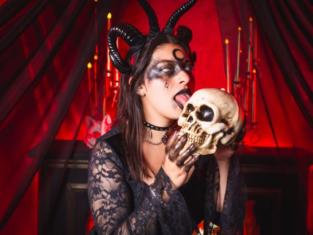 IsabellaFleur with black lace top, horned headpiece, and skull in hand, tongue out, red eyes, dark makeup, black choker, licking skull, red and black candle background.
