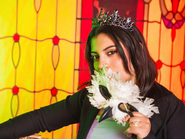 IsabellaFleur with dark hair, wearing a silver tiara, black top, holding a white feathered fan, looking at camera with sultry eyes, yellow and red stained glass background.