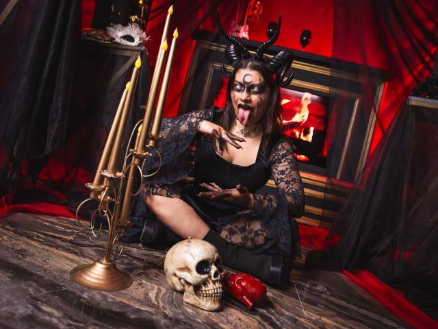 IsabellaFleur with black lace dress, devil horns, and face paint, tongue out, kneeling with gold candles, skull, and red apple on a wooden floor, in a dark, red-lit room.