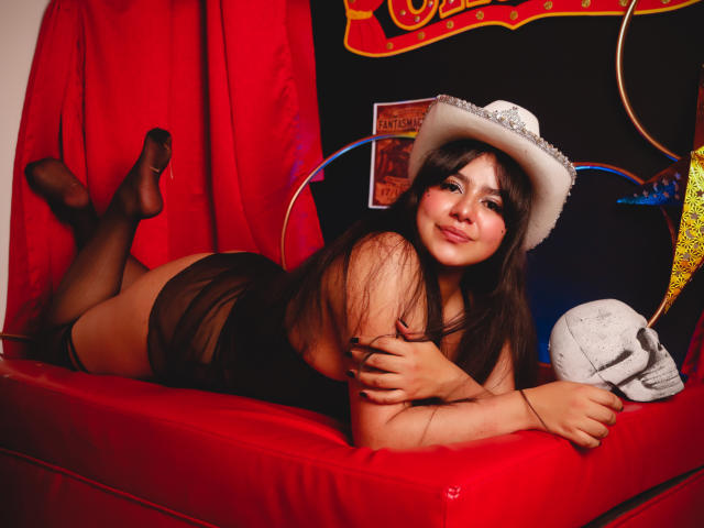 NicoleSamanta lying on a red couch, wearing a white cowboy hat, black sheer lingerie, and black stockings, with her ass up, smiling, holding a skull, and legs bent.