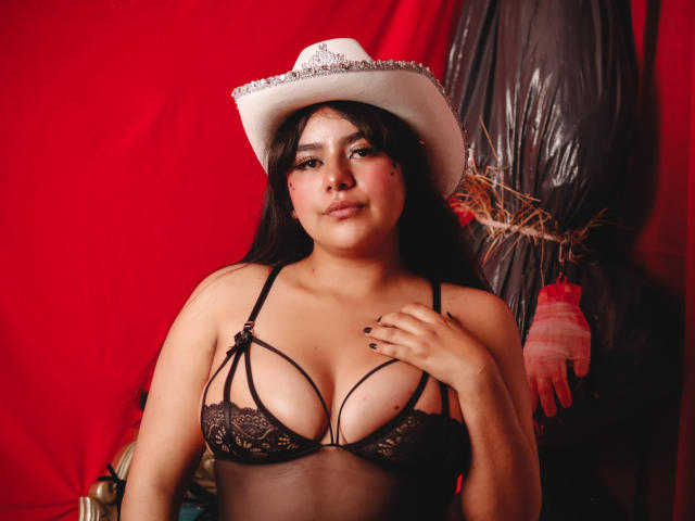 NicoleSamanta with dark hair, wearing a white cowboy hat and black lace bra, showing her tits, standing against a red backdrop with a plastic-covered object behind her.