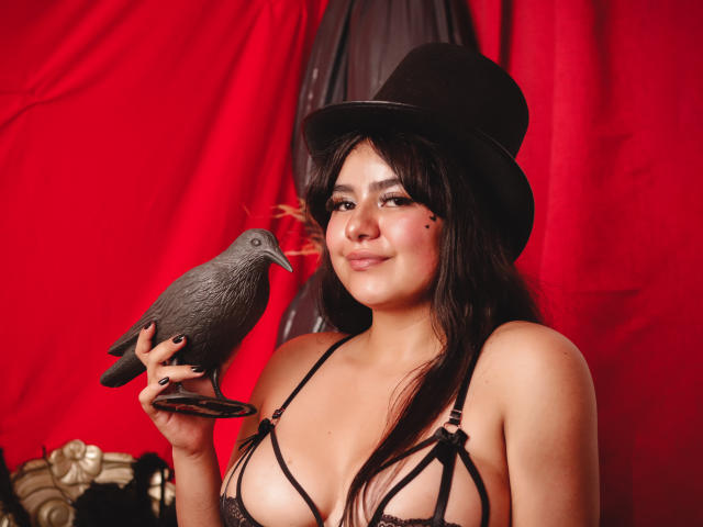 NicoleSamanta with dark hair, wearing a black top hat and strappy black lingerie, holding a grey pigeon, smiling, red backdrop, black nail polish, small black mole on her cheek.