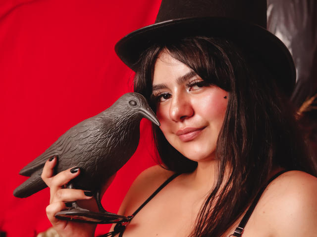 NicoleSamanta with dark hair, wearing a black top hat, holds a gray pigeon to her cheek, smiling, black nails, red background, and a small black mole on her right cheek.