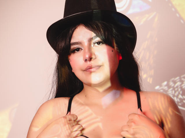 NicoleSamanta with long black hair, wearing a black hat and black spaghetti straps, holding her small tits with a slight smile, shadowed by light patterns on her face.