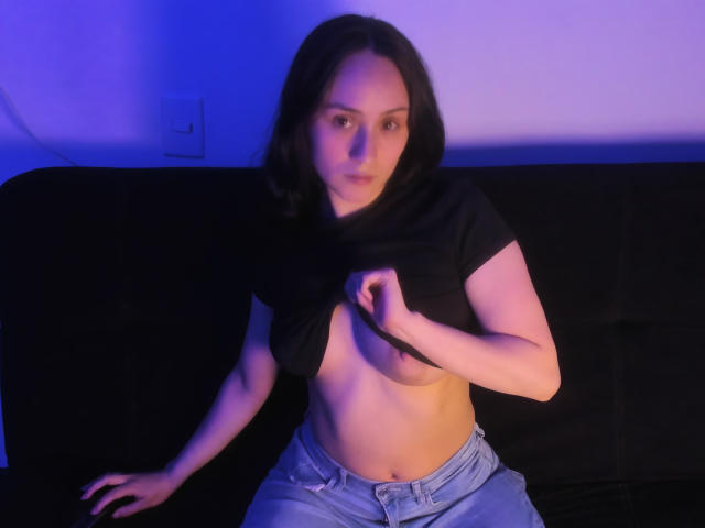 AnnieLamberth, with dark hair, lifting a black shirt to reveal her small, perky tits, sitting on a black couch, wearing light blue jeans, in blue and purple lighting.