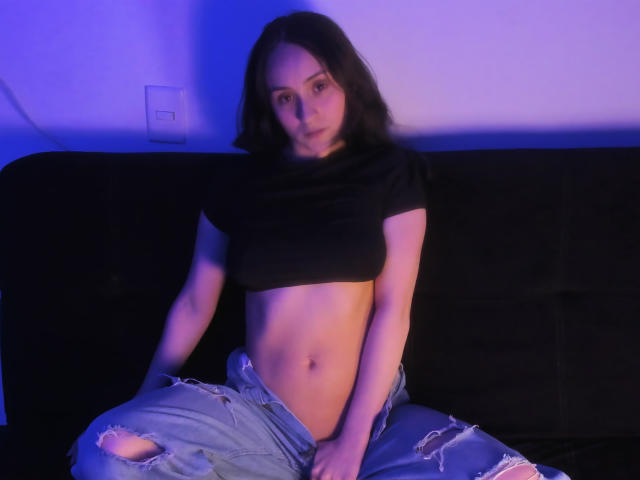 AnnieLamberth with dark hair, small tits, black crop top, and ripped jeans, sitting on a black couch, blue and purple lighting, looking at camera, hands on knees.