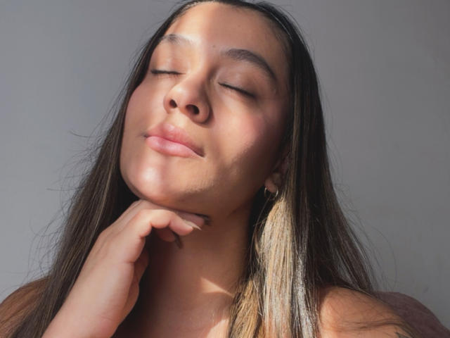 Thalia Ares tiene los ojos cerrados, un rostro iluminado por el sol, cabello castaño largo, labios ligeramente entreabiertos, y su mano tocando su barbilla, con una expresión suave y relajada..