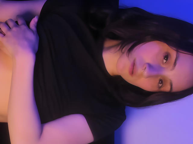 AnnieLamberth, with black hair and a nose ring, lying down, wearing a black shirt, hands on her stomach, in a blue and purple-lit image, showing her tits.