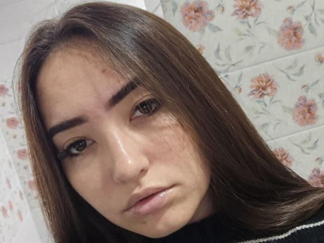 EmelineMinacucci has brown eyes, straight brown hair, and a small zit on her right cheek. Her eyebrows are thick, and she's wearing black eyeliner. Her lips are slightly parted.